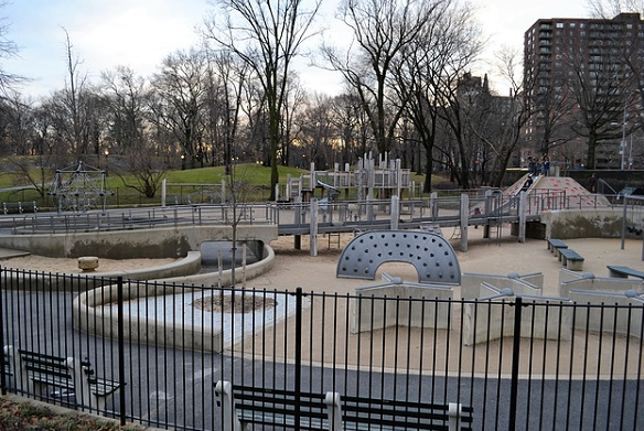 Bild 1: Vom Zaun umgebener Spielplatz mit Sitzbänken am Wegrand und auf dem Spielplatz