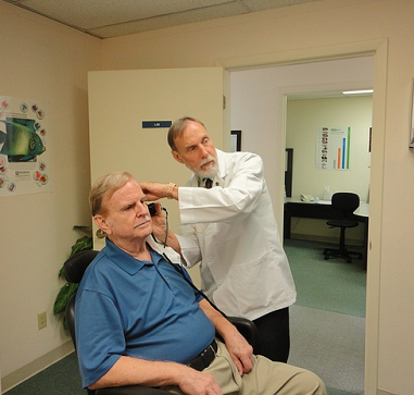 Bild 2: Hörakustiker führt bei älterem, rechts neben im sitzenden, Mann einen Hörtest durch